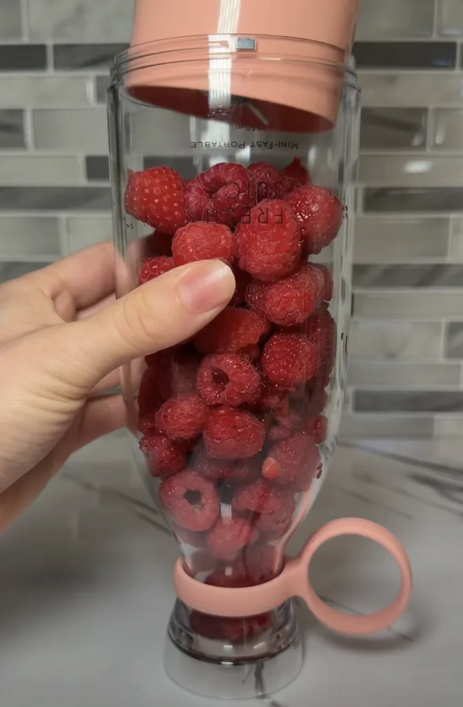 A clear portable blender bottle filled with vibrant red raspberries held by a hand on a clean kitchen countertop