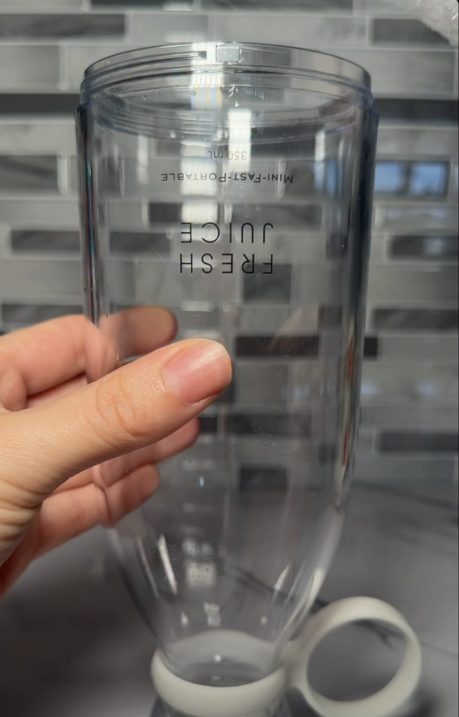 A person s hand gently holding a clear empty blender bottle on a white marble countertop Small water droplets are visibl