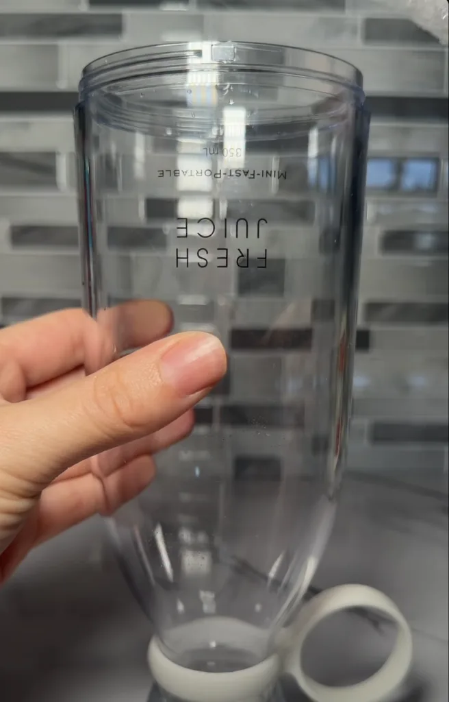 A person s hand gently holding a clear empty blender bottle on a white marble countertop Small water droplets are visibl