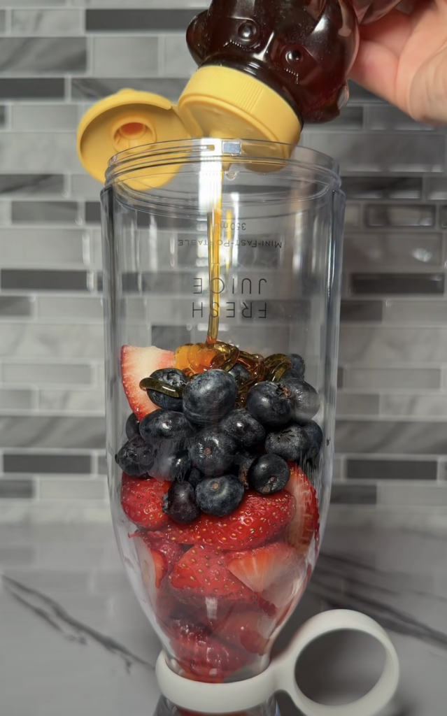 A clear blender cup filled with strawberries and blueberries with a hand pouring golden honey from a bear-shaped dispens