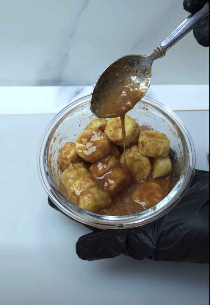 A clear glass bowl filled with golden-brown banana bites generously coated in a dark amber spiced caramel sauce A silver