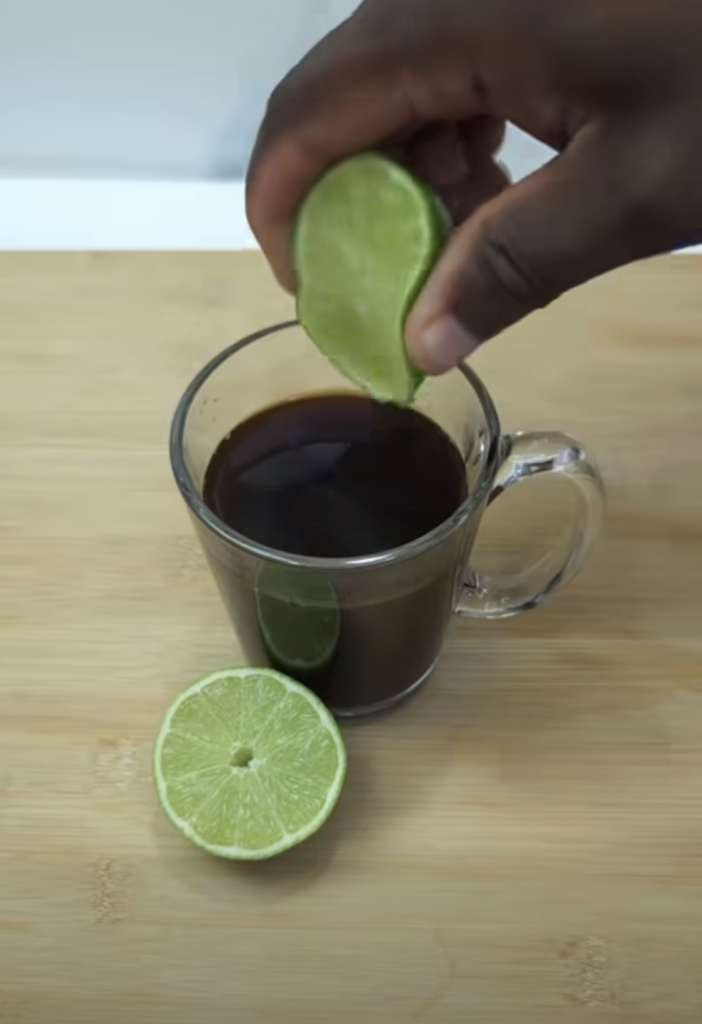 A hand squeezing a lime wedge into a mug of coffee on a light wooden surface