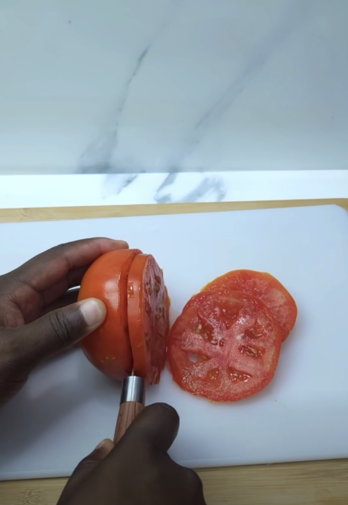 Dark-skinned hands slicing a red tomato on a white cutting board with a sharp knife alongside thin glistening tomato sli