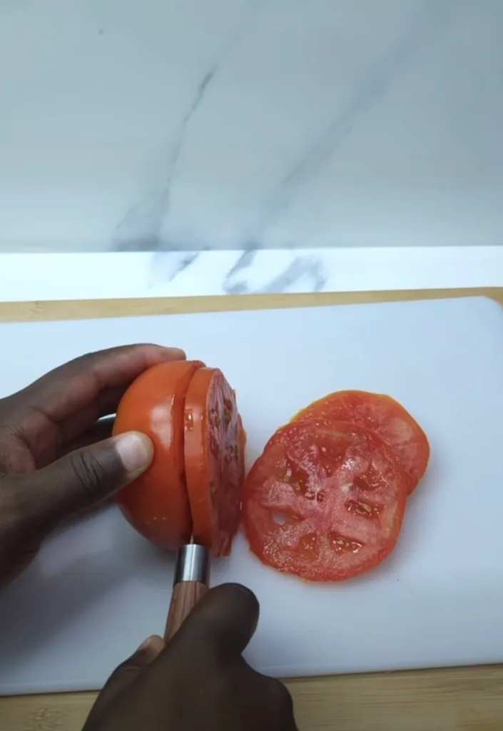 Dark-skinned hands slicing a red tomato on a white cutting board with a sharp knife alongside thin glistening tomato sli