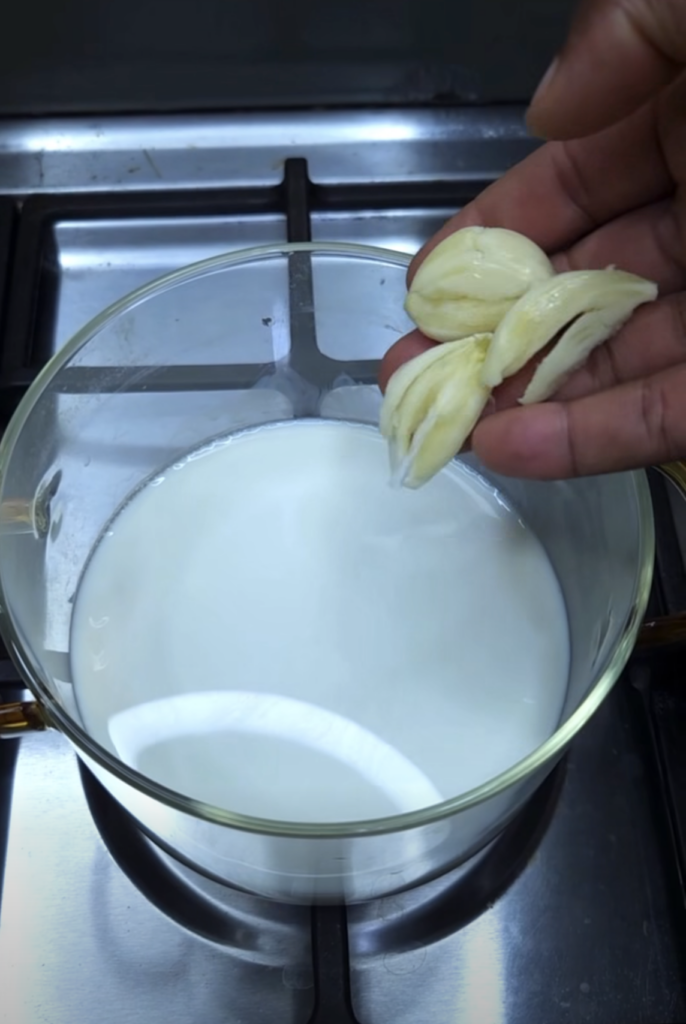 A hand holding several peeled garlic cloves poised above a clear glass pot filled with white milk on a modern gas stove 