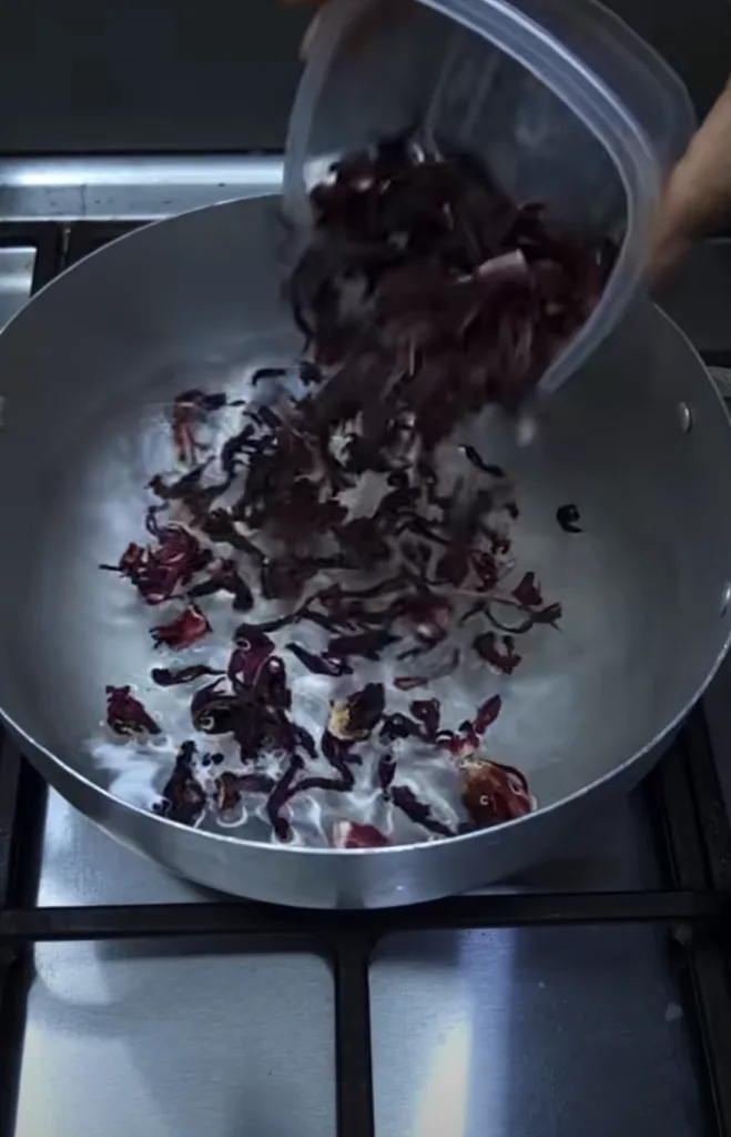 A close-up overhead view of a hand pouring dried dark red hibiscus petals into a metal pot filled with water with many p