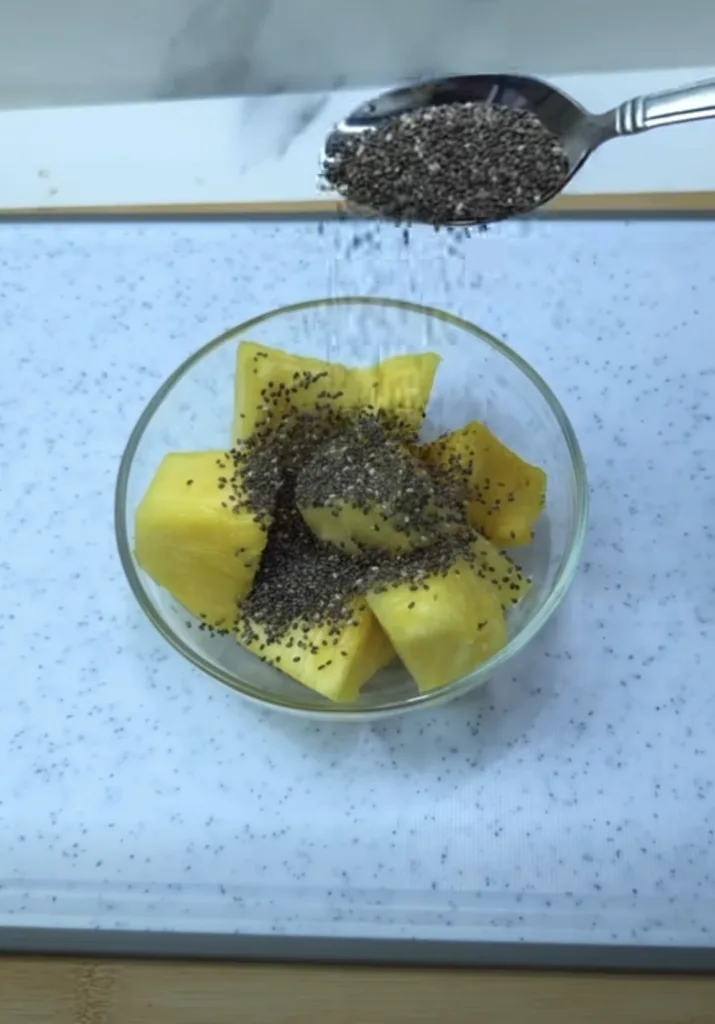 An overhead shot of a clear glass bowl filled with bright yellow pineapple chunks on a white speckled counter A silver s