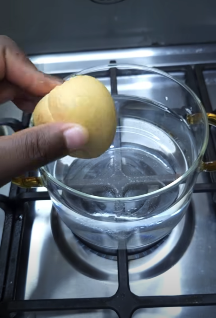 A dark-skinned hand holding a yellow lemon above a clear glass pot filled with water on a gas stove burner illustrating 