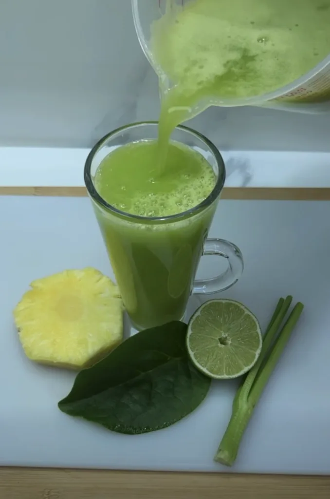 Vibrant green juice being poured from a clear measuring cup into a tall glass mug surrounded by fresh pineapple lime and