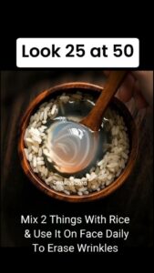 A bowl of creamy rice paste with a wooden spoon surrounded by grains of uncooked rice and a small bottle of milk or rose