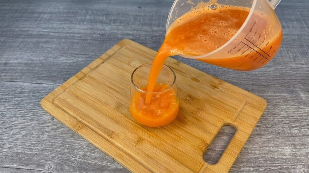 Vibrant orange juice being poured from a clear pitcher into a glass tumbler on a wooden cutting board