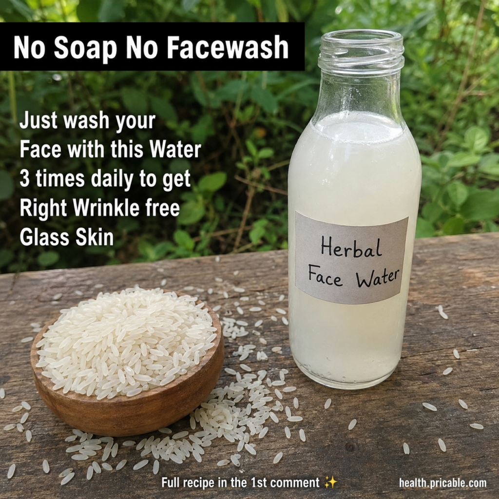 A clear glass bottle of milky rice water labeled Herbal Face Water next to a wooden bowl of uncooked white rice on a rus