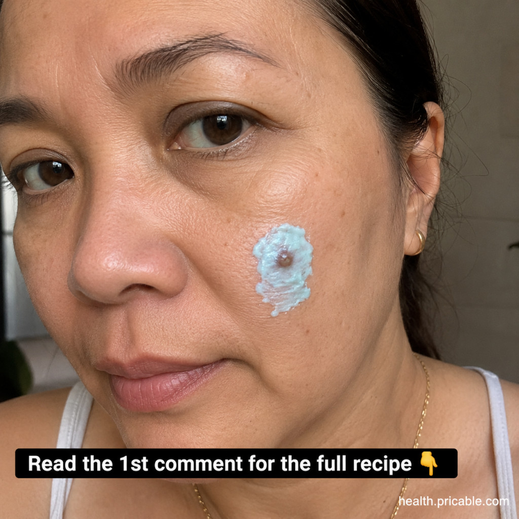 A woman applies a light-colored paste to a small skin imperfection on her cheek looking directly at the camera The paste