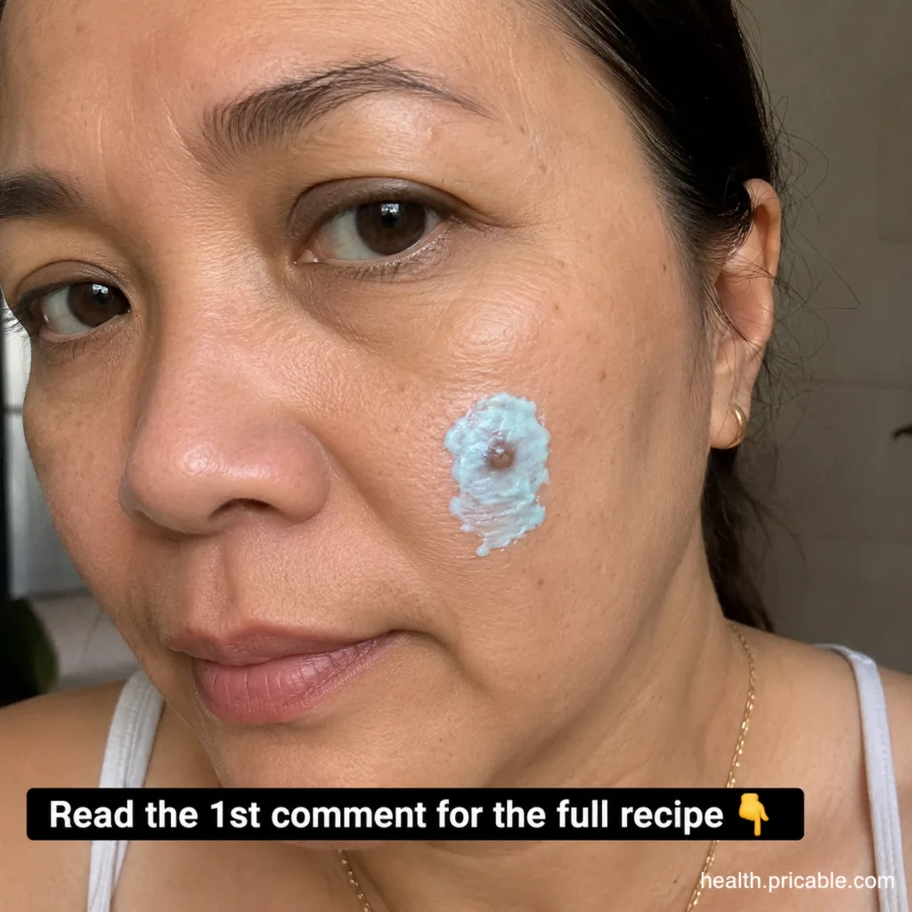 A woman applies a light-colored paste to a small skin imperfection on her cheek looking directly at the camera The paste