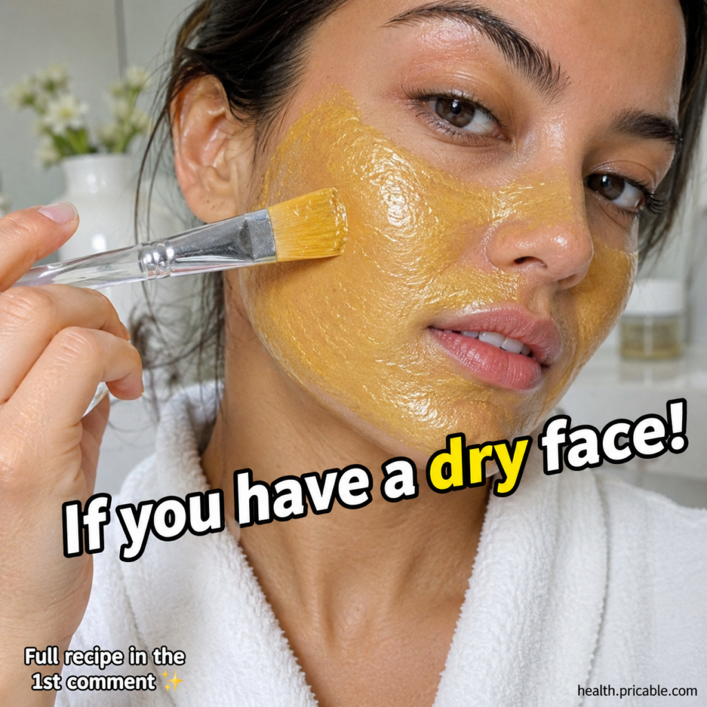 A woman applying a golden-yellow face mask to her cheek with a clear applicator brush looking directly at the viewer She