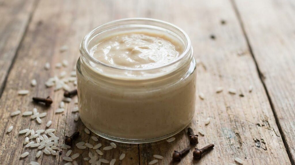 A small glass jar filled with a light creamy homemade collagen cream next to fresh rice grains and dried cloves