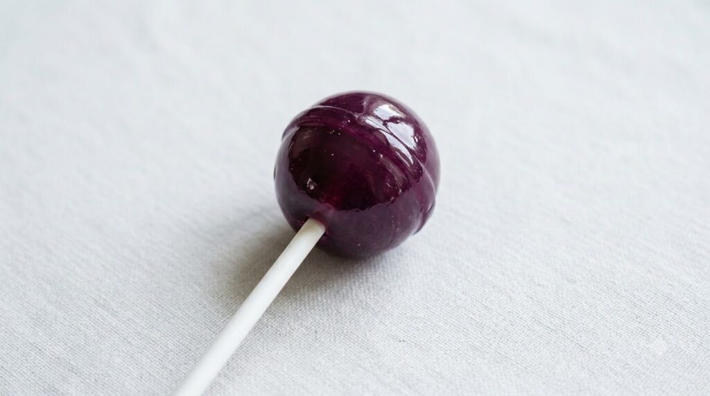 A deep purple grape-flavored fruit lollipop with a stick smooth and glossy on a light background