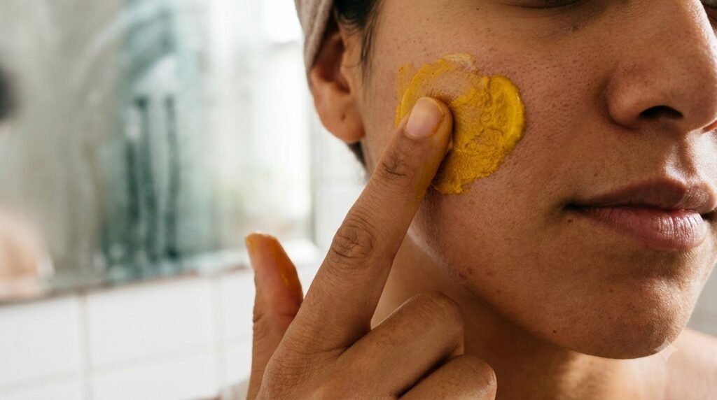 Close-up of a hand gently applying a yellow turmeric and Vaseline paste to a cheek with a blurred background of a bathro