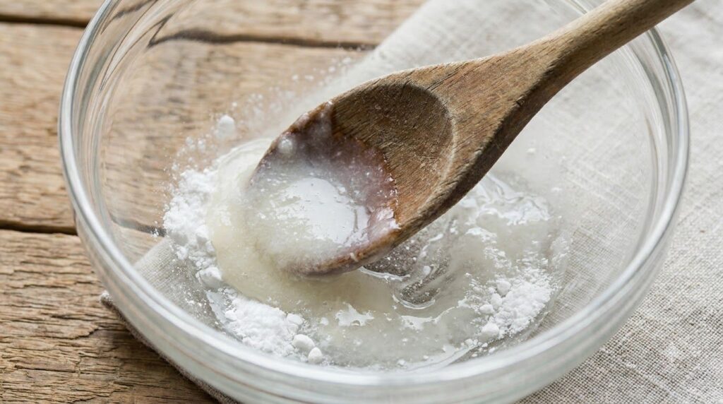 A close-up of crushed white aspirin powder being mixed with clear baby oil in a small glass bowl to form a paste