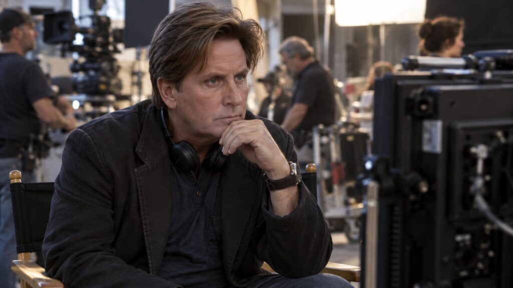 Emilio Estevez in a director s chair looking intently at a monitor on a film set with crew members in the background con