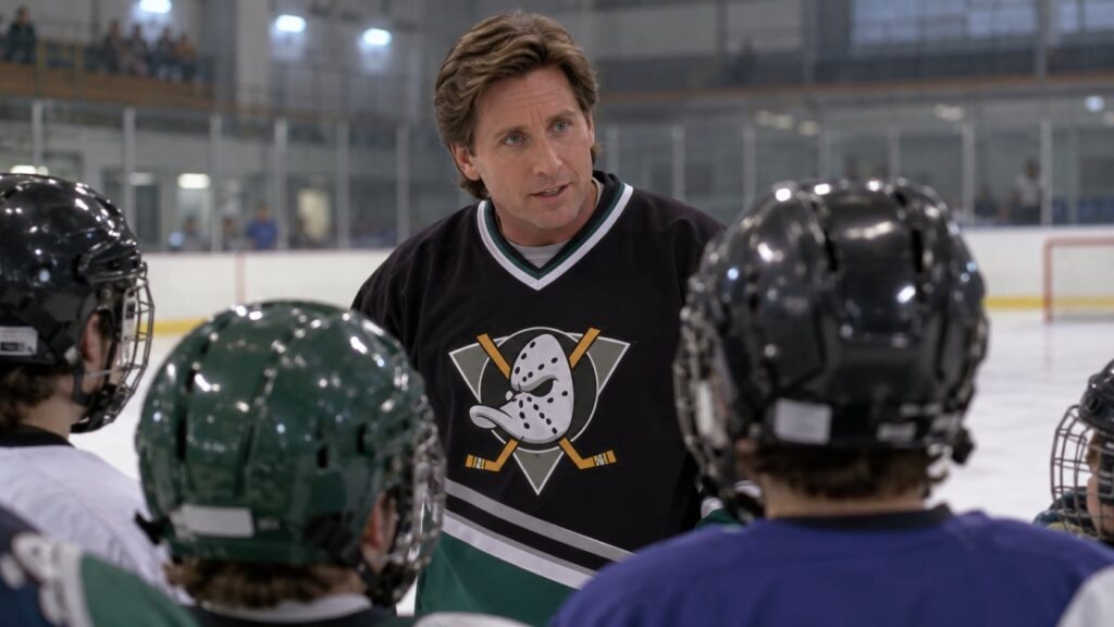 A still from The Mighty Ducks showing Emilio Estevez as Coach Gordon Bombay on an ice rink surrounded by young hockey pl