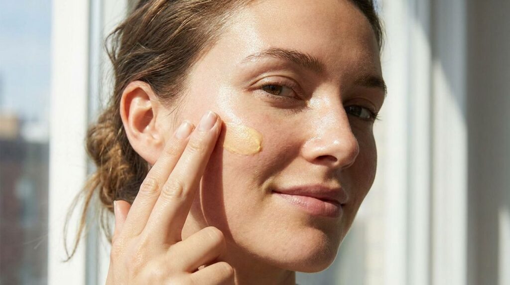 A woman gently massaging a small amount of yellow cream onto her cheek emphasizing clean glowing skin