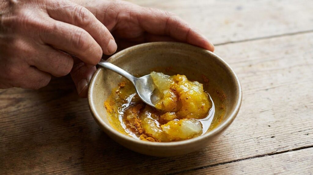 A hand mixing petroleum jelly glycerin and turmeric powder in a small bowl with a spoon showing the consistency of the f