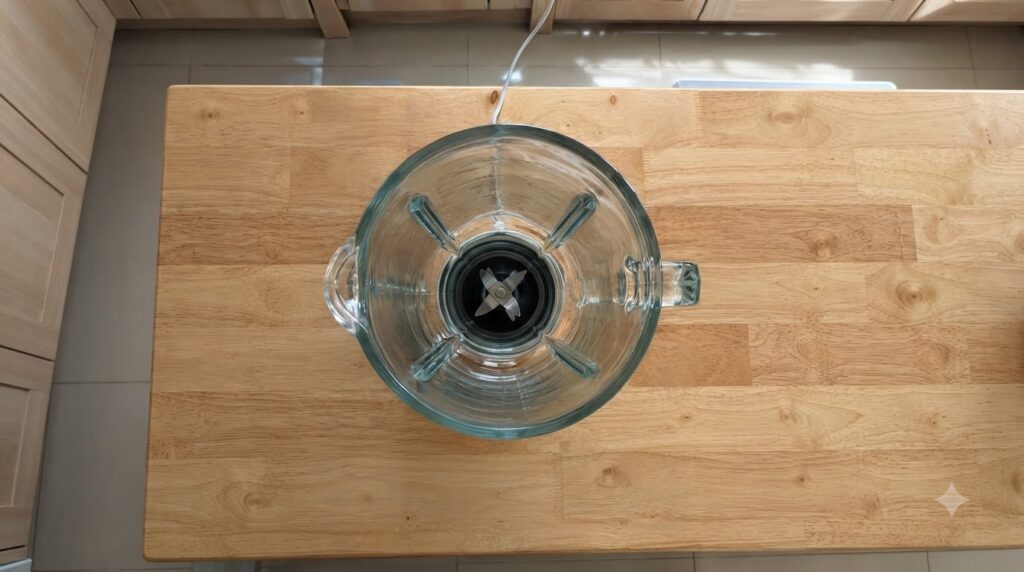 A clean empty glass blender pitcher with sharp metallic blades resting on a warm wooden countertop with a white power co