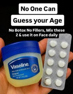 A small clear glass jar filled with a creamy glossy balm alongside an open Vitamin E capsule and a tub of Vaseline in th