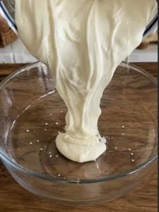 A thick off-white creamy substance being poured into a clear glass bowl showcasing its viscous texture and soft ripples 