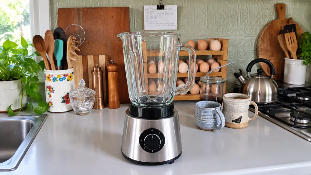 A stainless steel blender with a clear glass jar on a white countertop in a rustic kitchen setting surrounded by various