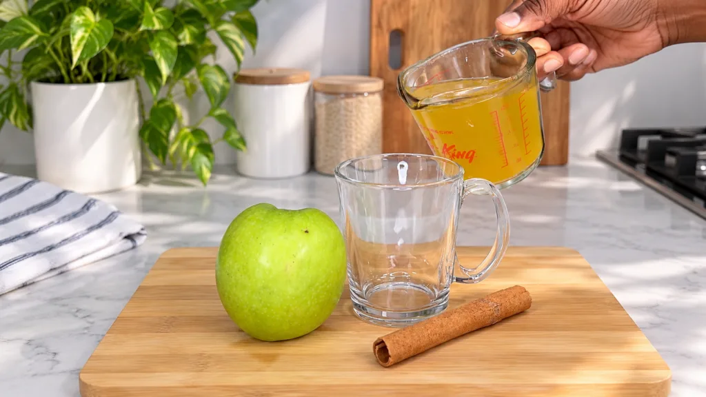 A person s hand is actively pouring a vibrant yellow liquid from a clear measuring cup into an empty clear glass mug On 