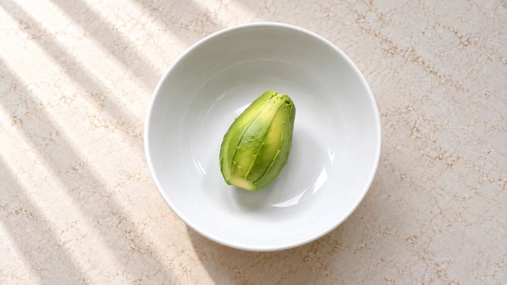 A perfectly peeled avocado half centered within a pristine white ceramic bowl set against a light beige mottled countert