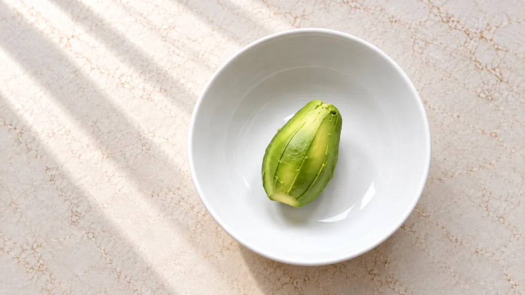 A perfectly peeled half avocado pit removed in a minimalist white ceramic bowl on a light marble surface with soft natur