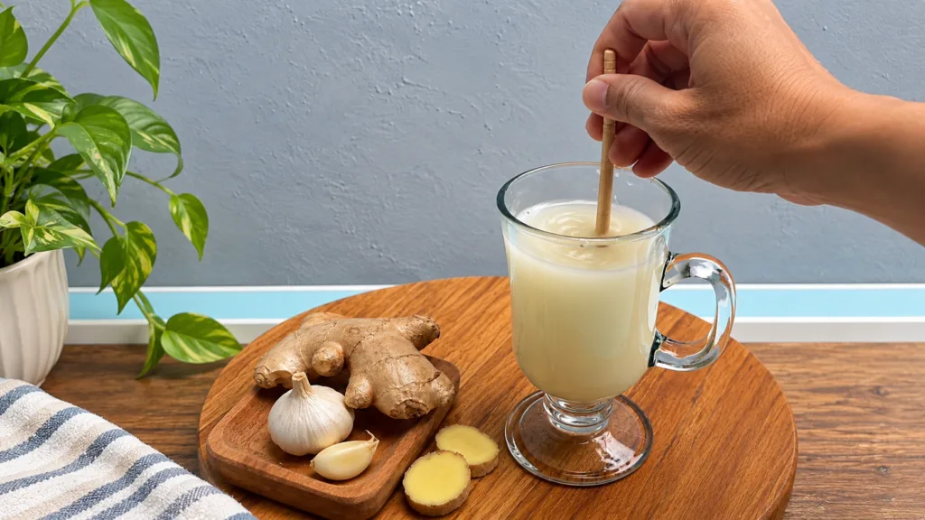 A close-up shot of fresh ginger roots and garlic cloves arranged on a wooden board next to a glass mug containing a ligh