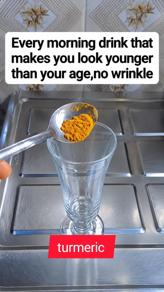 A hand holding a spoonful of bright yellow turmeric powder above an empty clear glass in a kitchen setting