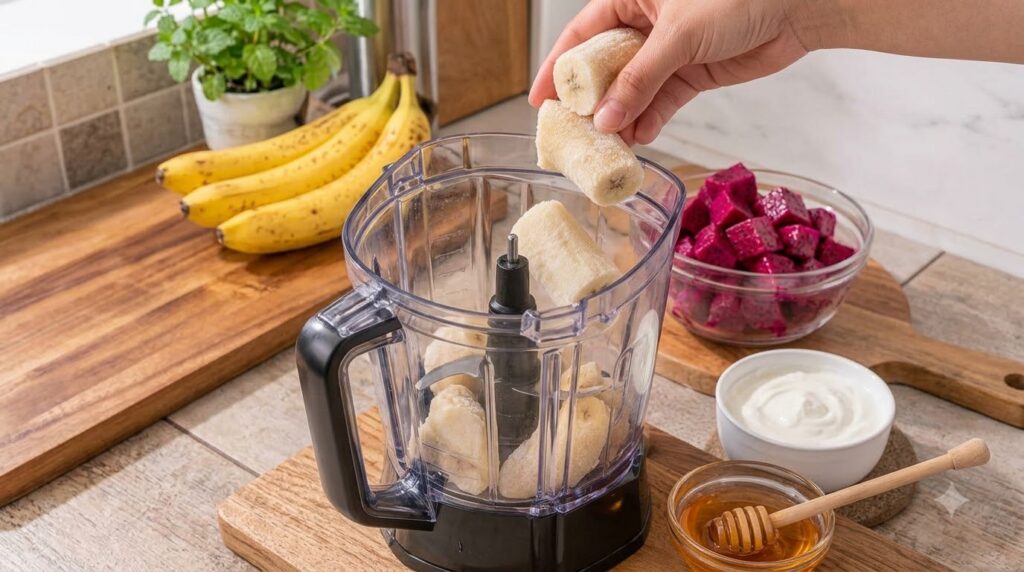 A top-down view of a hand dropping frozen banana slices into a blender surrounded by fresh bananas a bowl of diced drago