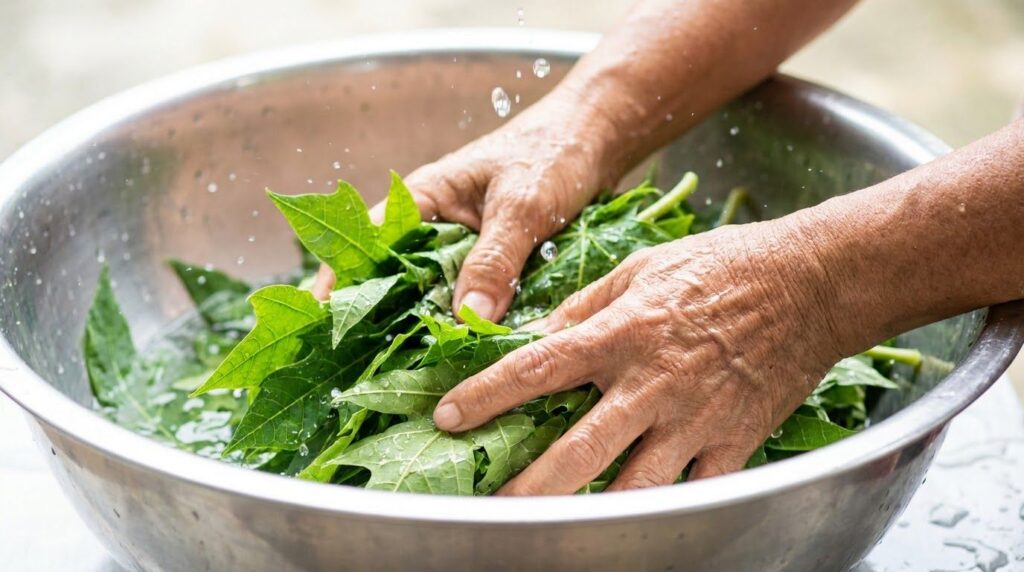 Hands washing a generous quantity of fresh green papaya leaves in a large metal bowl filled with water with visible wate