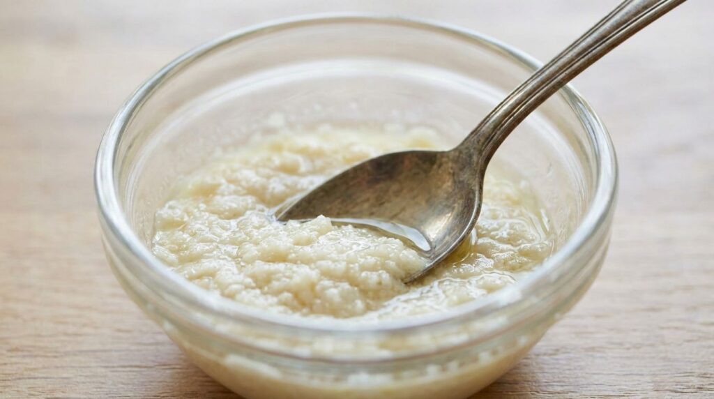 A close-up of a smooth white paste made from crushed aspirin and baby oil in a small glass bowl with a spoon resting in