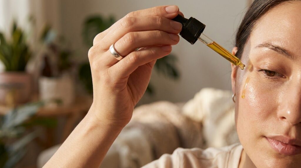 A person s hand applying a few drops of golden oil from a dropper bottle onto their clean face near the cheekbone area a
