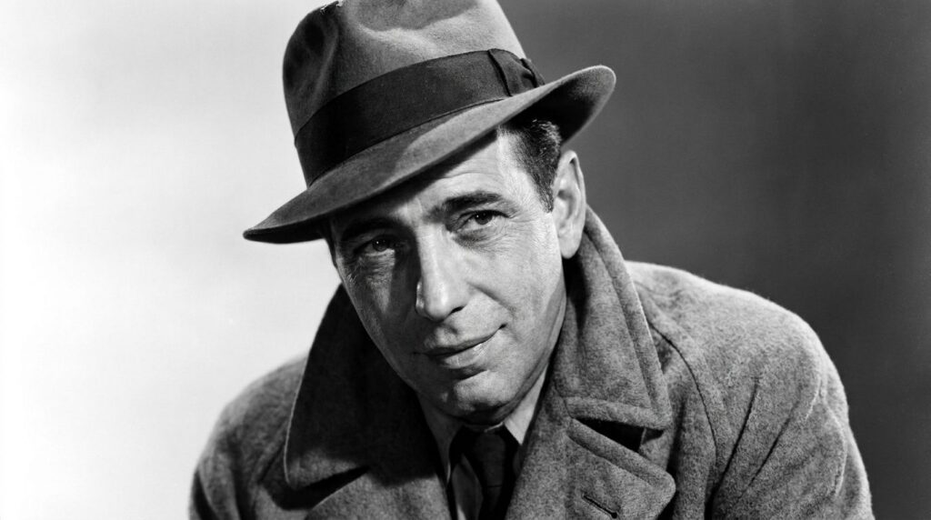 A black and white portrait of Humphrey Bogart featuring his classic fedora and trench coat look with a slightly cynical 