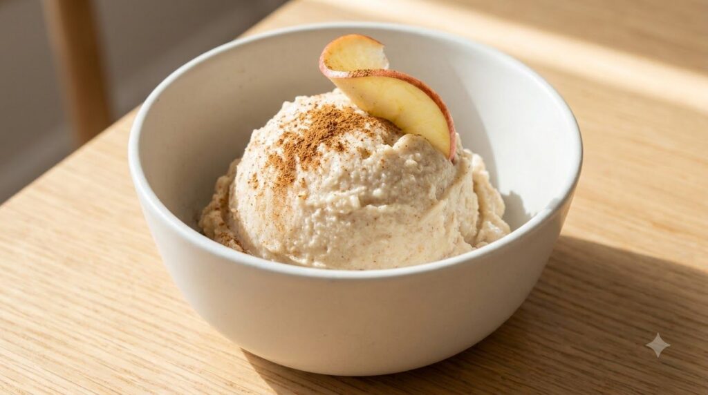 A creamy scoop of homemade apple pie nice cream in a white bowl topped with a dash of cinnamon and a small apple slice T