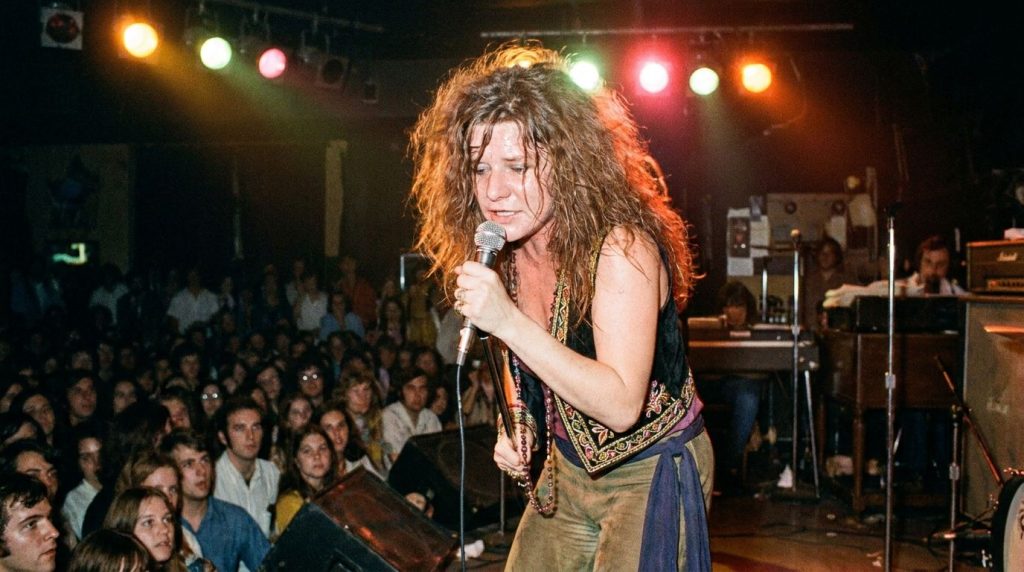 Janis Joplin performing live on stage with a microphone surrounded by band members radiating energy and passion in a vin