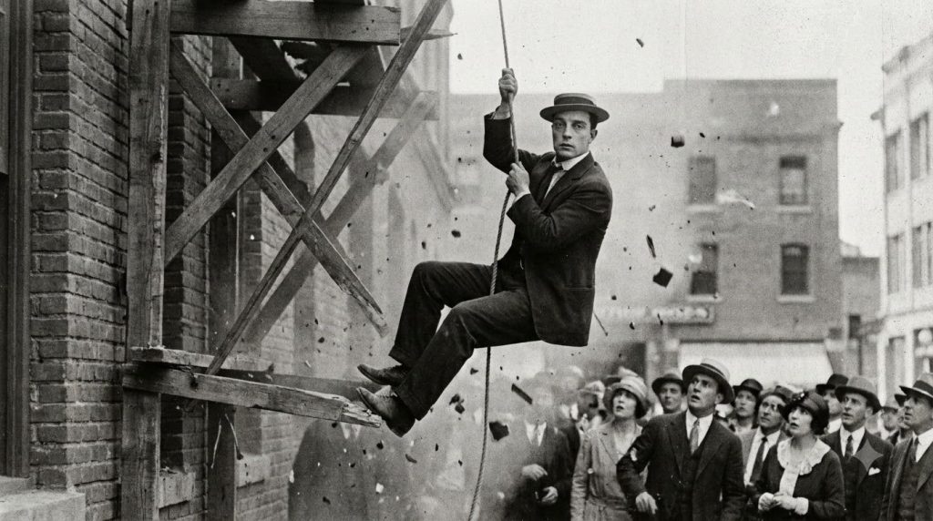 A black and white still from a Buster Keaton silent film showing him performing a physical stunt with a deadpan expressi