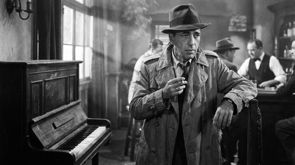 Humphrey Bogart as Rick Blaine in Casablanca leaning against a piano with a cigarette looking pensive and iconic