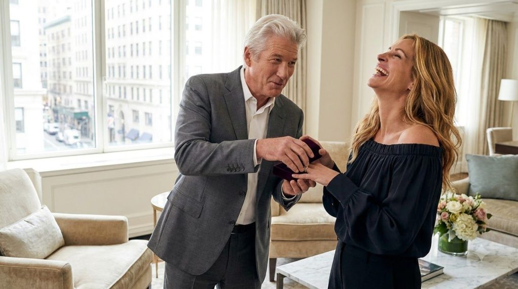 Richard Gere with a playful smile snapping a jewelry box shut in front of Julia Roberts who is laughing genuinely in a f