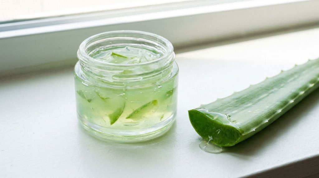 A small container of aloe vera infused Vaseline next to a fresh aloe vera leaf on a clean white background emphasizing n