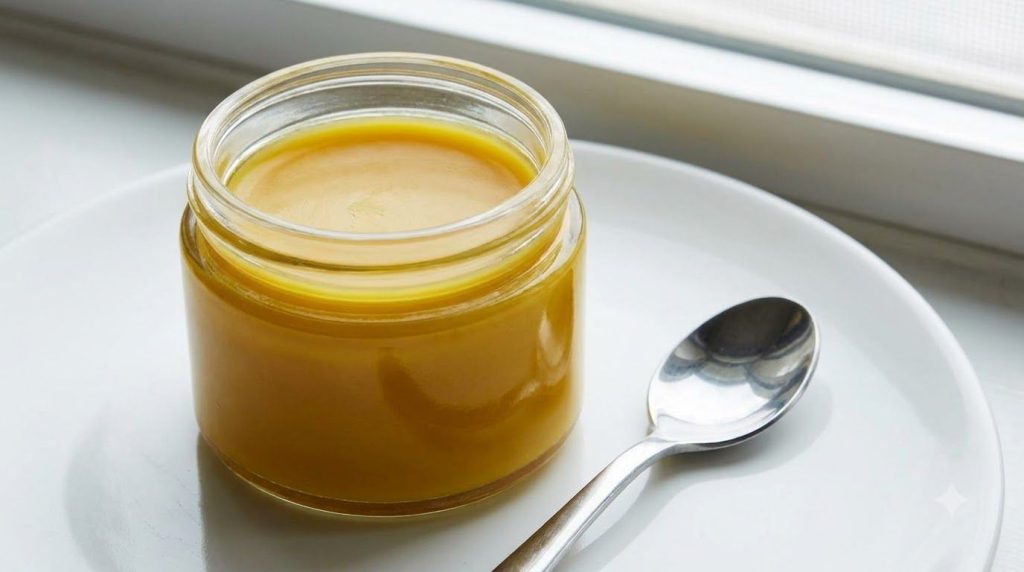 A close-up shot of a small glass jar filled with a golden turmeric and Vaseline mixture with a spoon resting beside it o