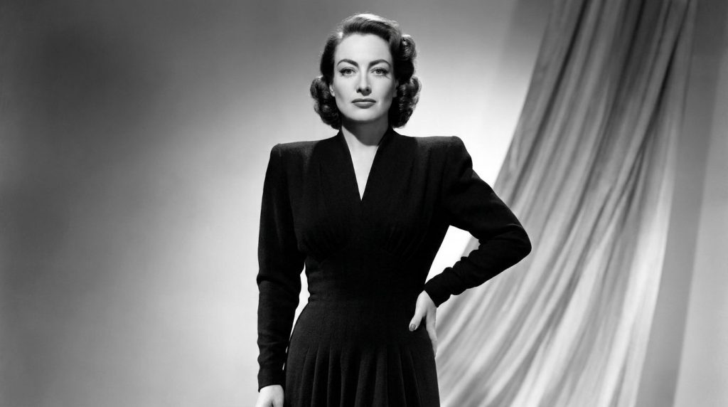 A black and white photograph of Joan Crawford in a stylish gown showcasing her signature broad-shouldered fashion and el