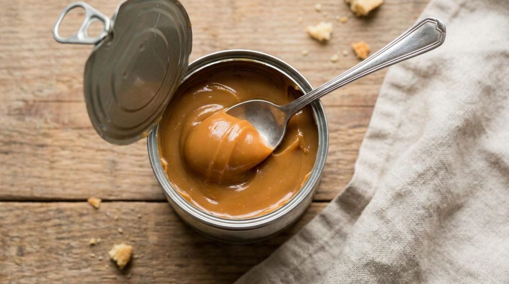 An opened can of homemade dulce de leche with a spoon scooping out creamy caramel